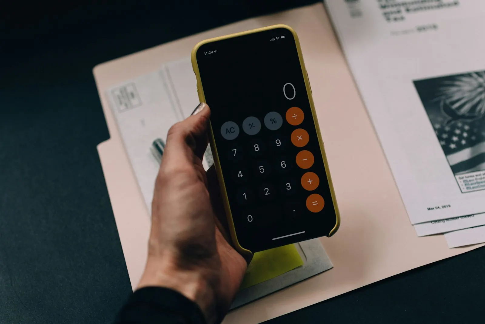 Mão a segurar um telemóvel com a aplicação de calculadora aberta, sobre documentos numa mesa escura